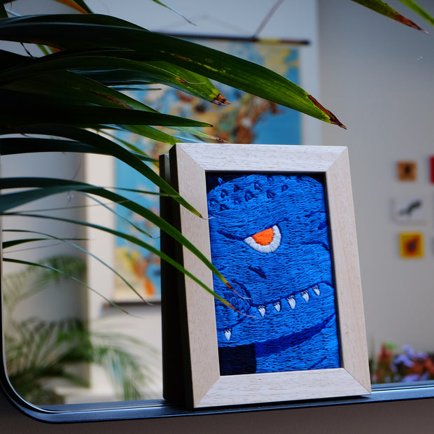 Alt angle of an embroidery piece in a thick light stained wooden frame. Embroidery is a close up shot of a deep blue Godzilla, with a single orange eye looking at the viewer menacingly and white teeth showing out of a closed mouth. Frame is propped on a mirror, slightly obstructed by a plant.