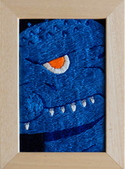 Embroidery piece in a thick light stained wooden frame. Embroidery is a close up shot of a deep blue Godzilla, with a single orange eye looking at the viewer menacingly and white teeth showing out of a closed mouth.