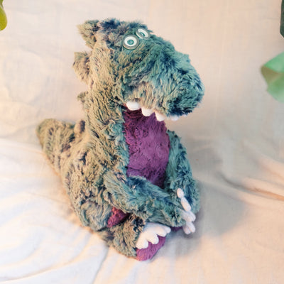 Soft plush sculpture of a timid looking Godzilla figure, with green fur and spikes and a purple belly. Plush has button eyes, white teeth and white claws.