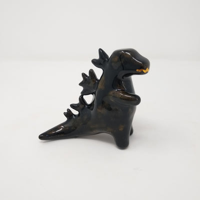 A ceramic black Godzilla figure, with minimal body and facial details. It has abstracted spikes on its back and a drawn on gold mouth.