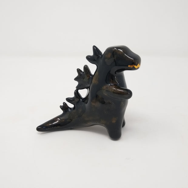 A ceramic black Godzilla figure, with minimal body and facial details. It has abstracted spikes on its back and a drawn on gold mouth.