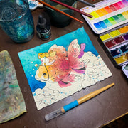 Watercolor painting on deckled edge paper of a nude woman with long blonde hair riding atop of a very large goldfish. They are against a teal blue background and above fluffy clouds. Piece is shown on a desk with watercolor, ink and various art tools.