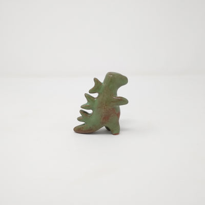 A small ceramic green and brown Godzilla figure, with no facial features and minimal body details. It has abstract spikes on its back.