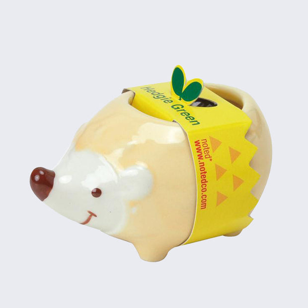 Small ceramic cartoon hedgehog, smiling with a brown nose. Its body is yellow and is wrapped in a cardboard packaging sleeve that says "Hedgie Green."