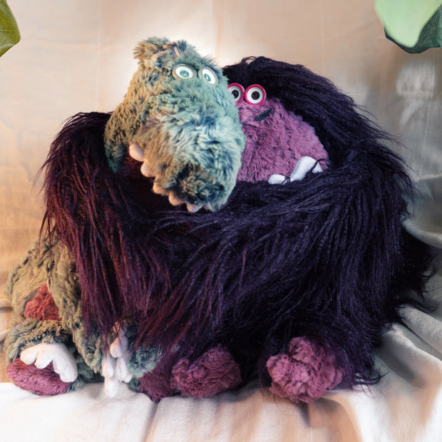 A purple plush King Kong with long fur and small button eyes wrapping its arms around a soft plush green Godzilla with pointy white teeth, small button eyes and white claws.