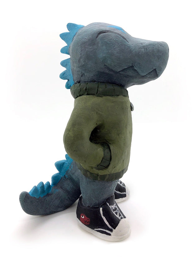 A painted clay sculpture of a happy looking grey blue Godzilla, with blue spikes and a long tail. He is wearing an army green jacket with a red dinosaur head logo and black sneakers. One hand is in his jacket pocket.