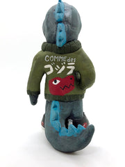 Back view of a painted clay sculpture of a happy looking grey blue Godzilla, with blue spikes and a long tail. He is wearing an army green jacket with a large red dinosaur head logo on the back of its jacket, which says "Comme des Gojira," with Gojira being written in Japanese.