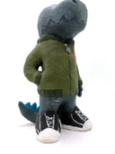 A painted clay sculpture of a happy looking grey blue Godzilla, with blue spikes and a long tail. He is wearing an army green jacket with a red dinosaur head logo and black sneakers. One hand is in his jacket pocket.