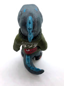 A painted clay sculpture of a happy looking grey blue Godzilla, with blue spikes and a long tail. He is wearing an army green jacket with a red dinosaur head logo and black sneakers. One hand is in his jacket pocket.
