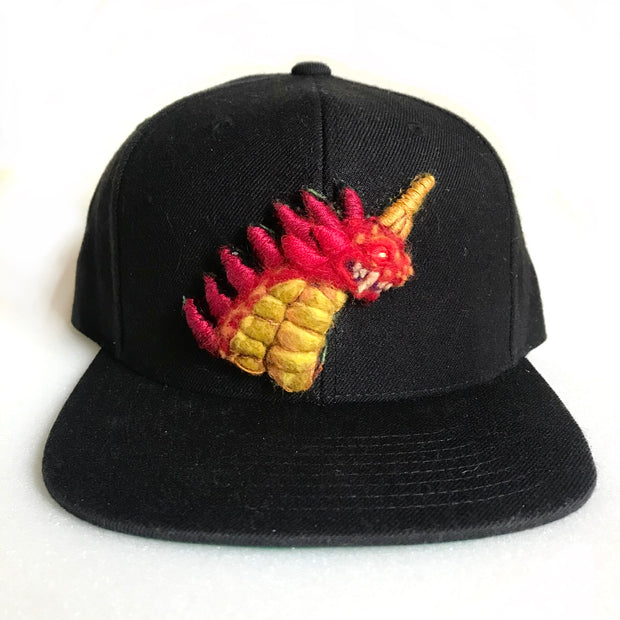 A black cap with a thickly felted Godzilla creature bust with red spikes, yellow body, glowing red eye and single yellow horn out of head.