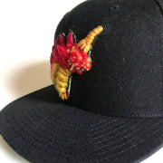 Side view of a black cap with a thickly felted Godzilla creature bust with red spikes, yellow body, glowing red eye and single yellow horn out of head.