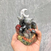 A hand holding  a ceramic sculpture of a two gray cats with green eyes emerging from a clustered city on fire. A crescent moon stands behind them.