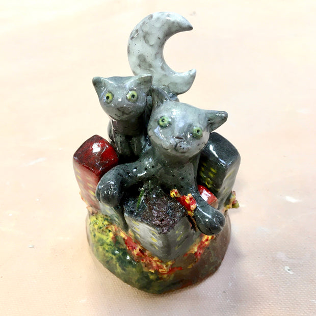 Ceramic sculpture of a two gray cats with green eyes emerging from a clustered city on fire. A crescent moon stands behind them.