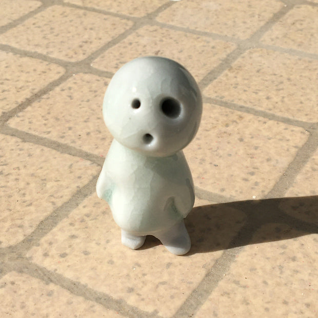 Small glazed porcelain sculpture of a Kodama, an off white forest spirit with simplistic body shape, round head and rounded arms and legs. It has asymmetrical eyes, both holes without any detailing and an "o" shaped small mouth. 
