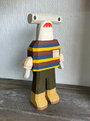 Sculpture made out of whittled wood and partially painted of a hammerhead shark wearing a striped shirt, pants and boots. The center of its body is very blocky.