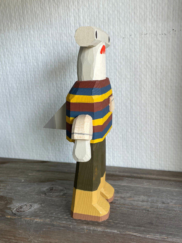 Sculpture made out of whittled wood and partially painted of a hammerhead shark wearing a striped shirt, pants and boots. The center of its body is very blocky and it has a pointed fin on its back.