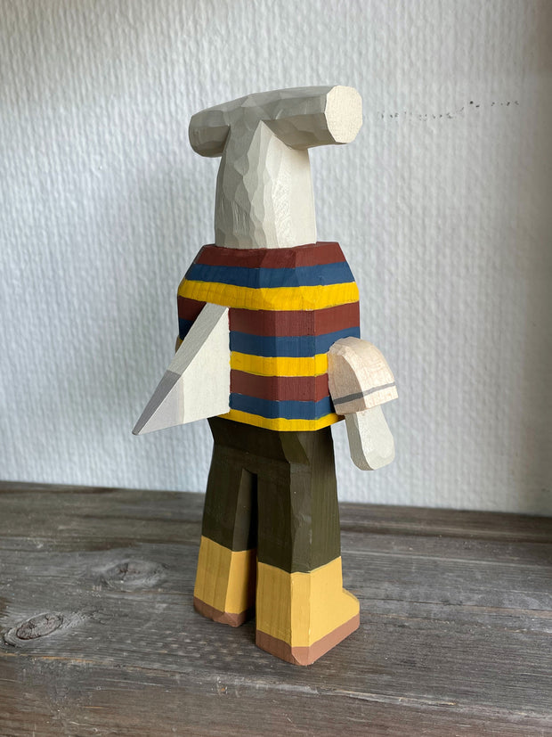 Sculpture made out of whittled wood and partially painted of a hammerhead shark wearing a striped shirt, pants and boots. The center of its body is very blocky and it has a pointed fin on its back.