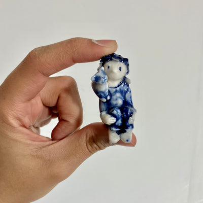 Small blue and white porcelain sculpture being held in someone's hand. Sculpture is of a small human-like figure sitting with their knees drawn into their chest with a small cat on its shoulder with its tail wrapped around the person's neck affectionately.
