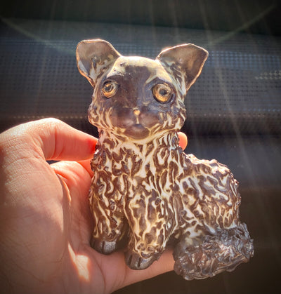 Brown and white ceramic sculpture of a cat, being held in someone's hand. Its eyes are painted gold and has characteristics of wild fur.