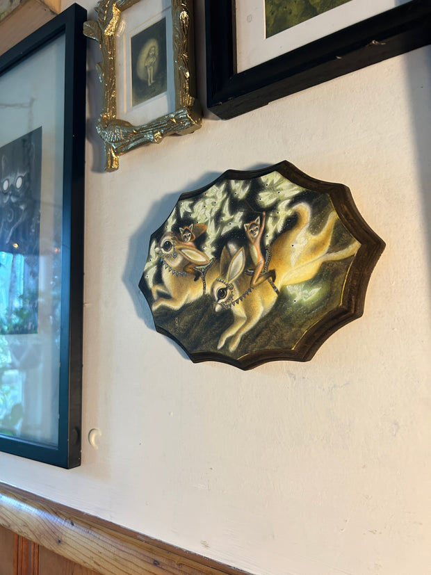 "The Rabbit Riders" being displayed on a white wall, slightly to the side to display the depth of the wooden plaque, about an inch deep. For description of piece, please read previous alt text.