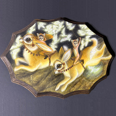 Illustration on a multi edged wooded plaque, of two large yellow rabbits with big brown eyes saddled and being rode by 2 small children in fox costumes, one holding a net up. Around them fly many illuminated white birds.