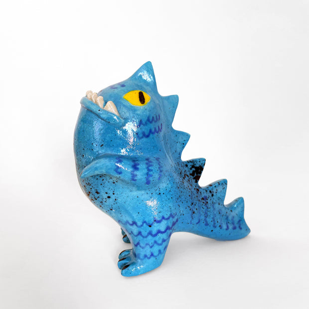 A ceramic sculpture of a chubby blue Godzilla-like figure, with yellow eyes and an underbite showing through closed mouth. Figure has blue drawn accents, black splatter accents and spikes along its back and tail.