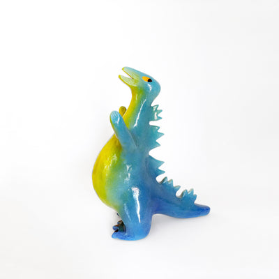 A ceramic sculpture of a pot bellied Godzilla-like figure, with a yellow stomach that fades into a green blue ombre across its body. It has yellow eyes and a slightly ajar mouth, with spikes along its back.