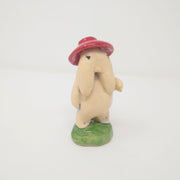 Ceramic sculpture of a cream colored creature with a face with two long cheek flaps, standing nude on a round green mound with a red hat.