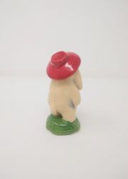 Ceramic sculpture of a cream colored creature with a face with two long cheek flaps, standing nude on a round green mound with a red hat.