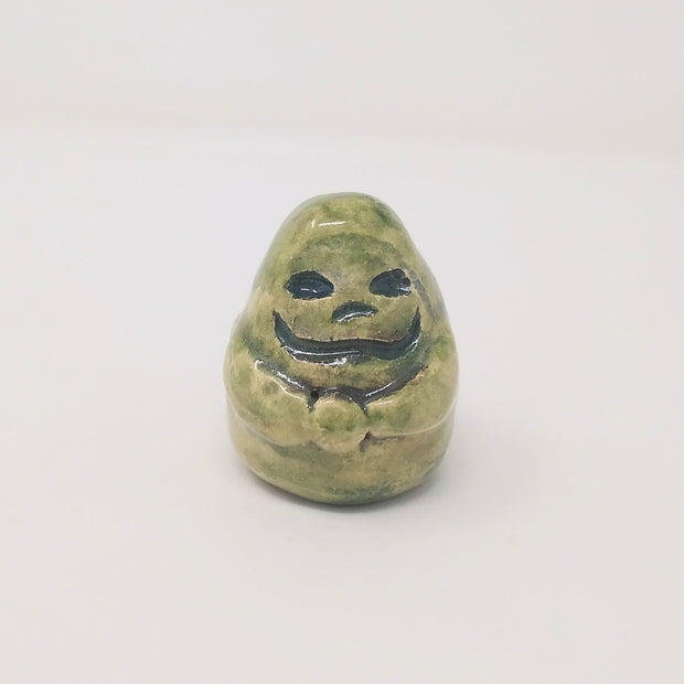 Ceramic sculpture of a small green, rounded old statue. It is green with a smile and simplified clasped hands. 