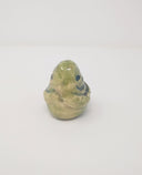 Ceramic sculpture of a small green, rounded old statue. It is green with a smile and simplified clasped hands.