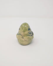 Ceramic sculpture of a small green, rounded old statue. It is green with a smile and simplified clasped hands.