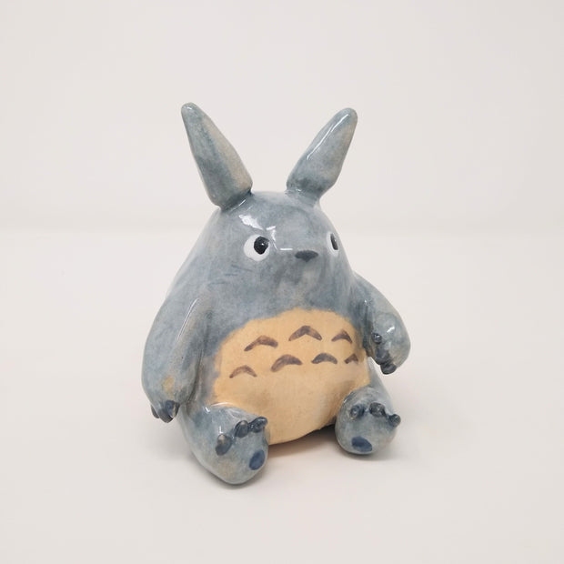 Glazed ceramic sculpture of Totoro, sitting and looking to the side curiously, with one paw slightly raised.