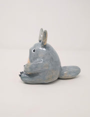 Glazed ceramic sculpture of Totoro, sitting and looking to the side curiously, with one paw slightly raised. He has a large, fluffy tail that lays flat behind him.