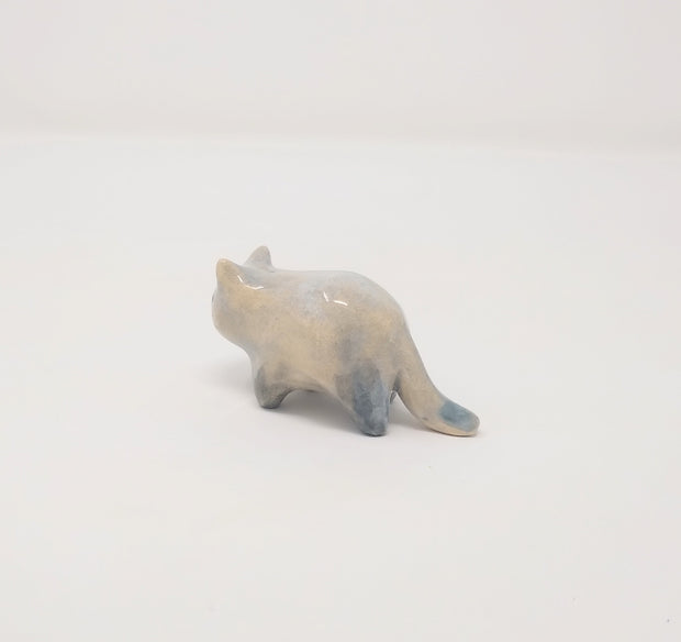 Small grey and white ceramic sculpture of a slightly chubby cat, standing on 4 legs with minimal shape details. It has a simplistic painted on cat face with whiskers.