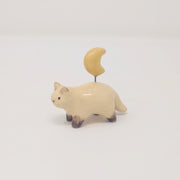 Small ceramic sculpture of a painted Siamese coloring cat, with simplistic body shapes and a drawn on face. Attached with a wire coming out of its back is a ceramic yellow crescent moon.
