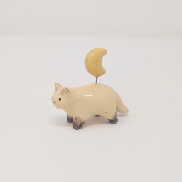 Small ceramic sculpture of a painted Siamese coloring cat, with simplistic body shapes and a drawn on face. Attached with a wire coming out of its back is a ceramic yellow crescent moon.