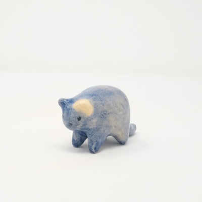 Small blue ceramic sculpture of a chubby blue cat with one cream colored ears. Body definition is minimal and has a small simple painted on cat face with whiskers.