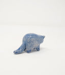 Small blue ceramic sculpture of a chubby blue cat with one cream colored ears. Body definition is minimal and has a small simple painted on cat face with whiskers.