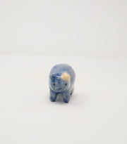 Small blue ceramic sculpture of a chubby blue cat with one cream colored ears. Body definition is minimal and has a small simple painted on cat face with whiskers.