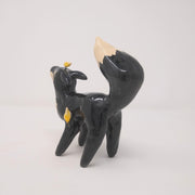 Ceramic sculpture of a black fox with white underbelly and tip of tail. It has non defined facial features and has a moon atop its head. Back view.