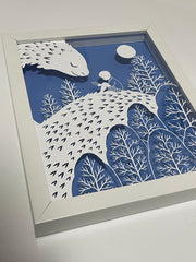 Side angle of a white cut paper illustration on a blue background in a white wooden frame. Cut paper design depicts a young child reading a book, sitting on a kind looking dragon, whose face is turned smiling to the child. A moon and trees are around the two.