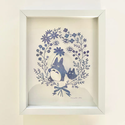 Delicate hand cut paper sculpture, of a small Totoro holding hands with an even smaller Totoro, they are framed in a delicate series of flowers and leaves. Framed in a white wooden frame.