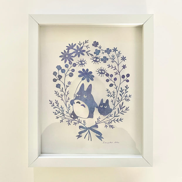 Delicate hand cut paper sculpture, of a small Totoro holding hands with an even smaller Totoro, they are framed in a delicate series of flowers and leaves. Framed in a white wooden frame.