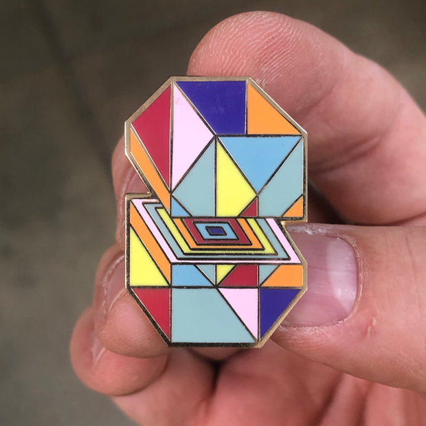 Enamel pin of an octagon filled with multicolored triangles, split in half to show rainbow squares stacked into one another. 