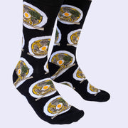 Side view of a model's legs wearing black socks. They have a ramen bowl pattern on them. The bowl is white and the broth inside is orange.