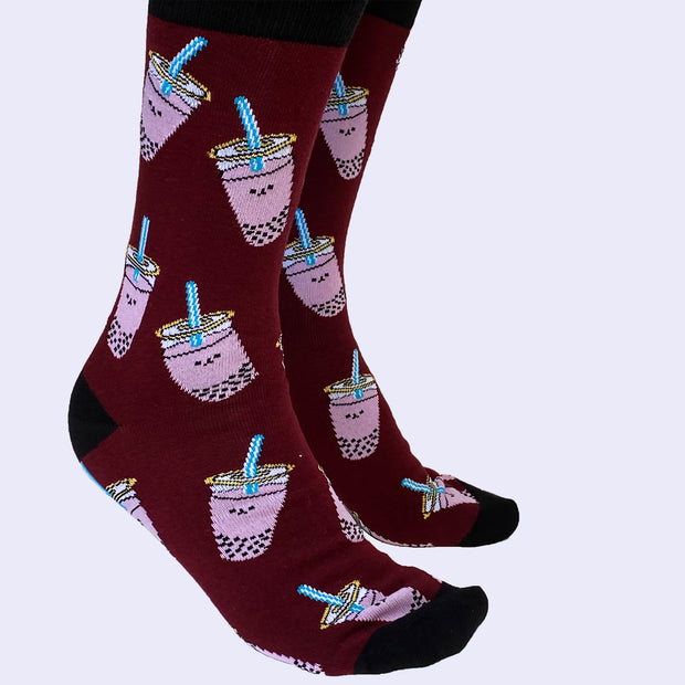 Side view of a model's feet wearing cartoon boba socks. The smiling purple cups are filled with black boba balls on the bottom.