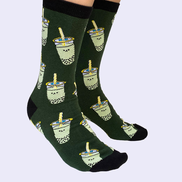Side view of model's feet wearing cartoon boba socks. The sock is dark green but the bobas are light green. The cuff, heel, and toe area is black