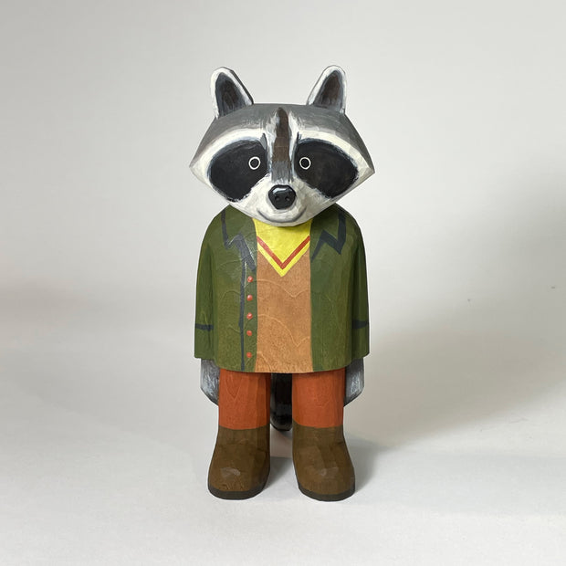 Small painted wooden sculpture of a sweet looking raccoon, standing and wearing a green coat, yellow and brown shirt, burnt orange pants and brown boots.