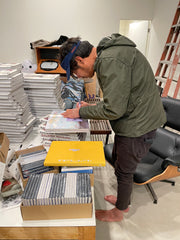 Barry McGee artist wearing green jacket is hunched over a stack of books and is signing one. You can't see what he's writing.